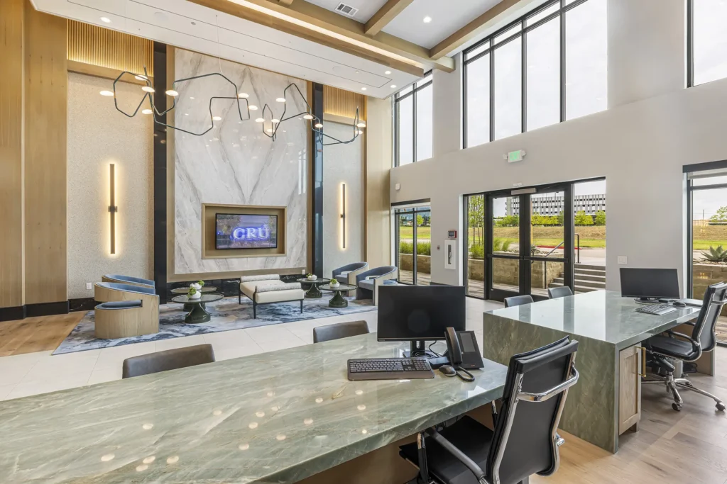 Modern office reception area with marble desk, computers, black chairs, stylish lighting, and a lounge space featuring a large TV, blue chairs, and decorative wall art. Floor-to-ceiling windows provide natural light.