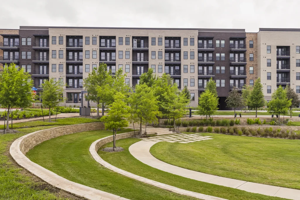 A modern apartment building with balconies stands behind a landscaped green area featuring curved sidewalks, young trees, trimmed grass, and low stone retaining walls.