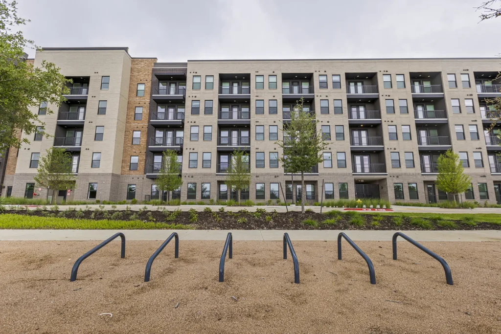 A modern, four-story apartment building with balconies is seen behind a row of small trees and a bike rack on a sandy area in the foreground. The sky is overcast.