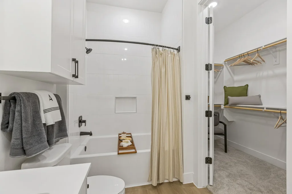 Modern bathroom with white walls, a bathtub with a beige shower curtain, a wooden bath tray, folded towels, and an adjacent walk-in closet featuring shelves, wooden hangers, and a green-cushioned bench.