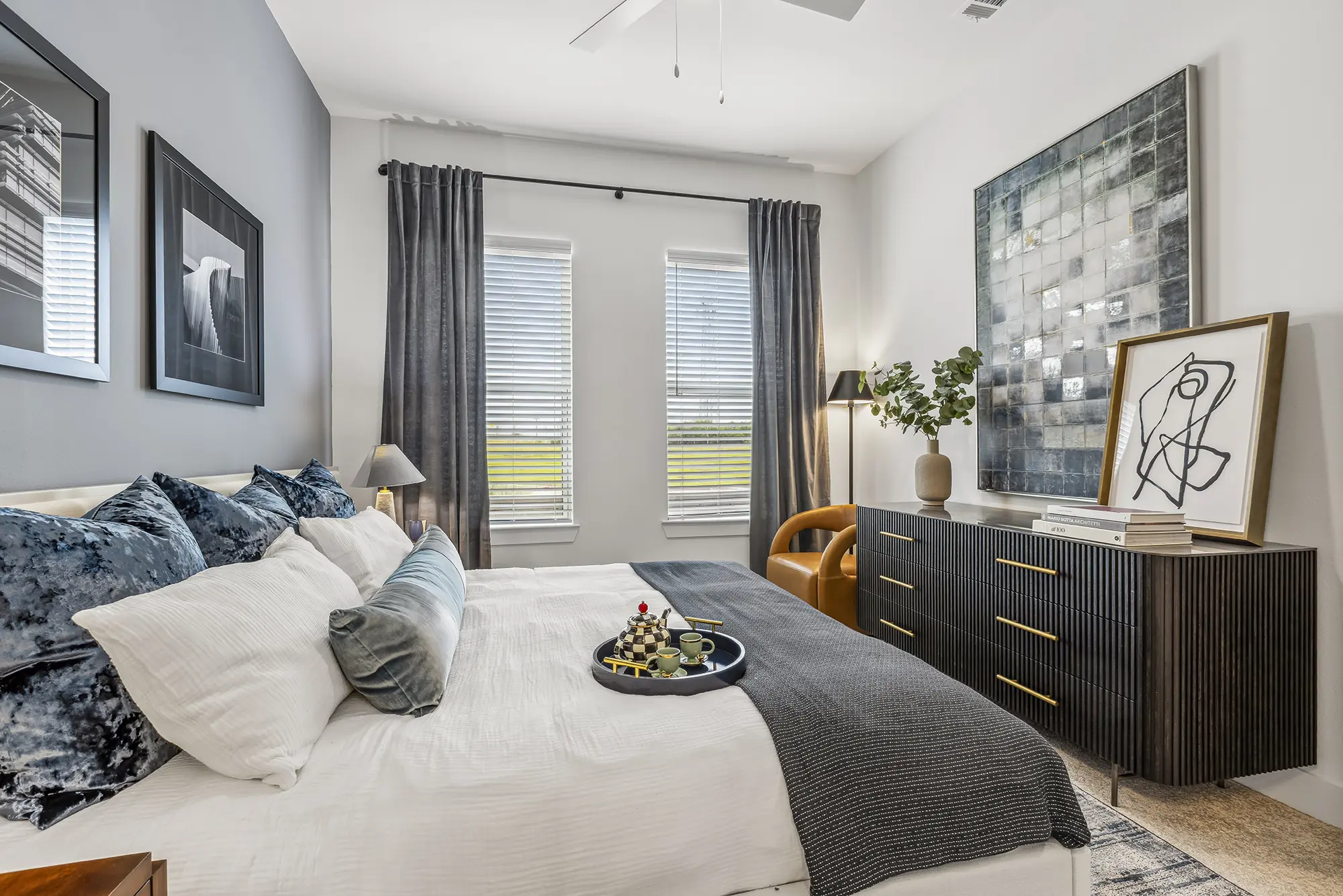 A modern bedroom with a neatly made bed, gray and white bedding, a tray with chess pieces, large windows with dark curtains, abstract art on the walls, a dark dresser, a brown chair, and a plant.