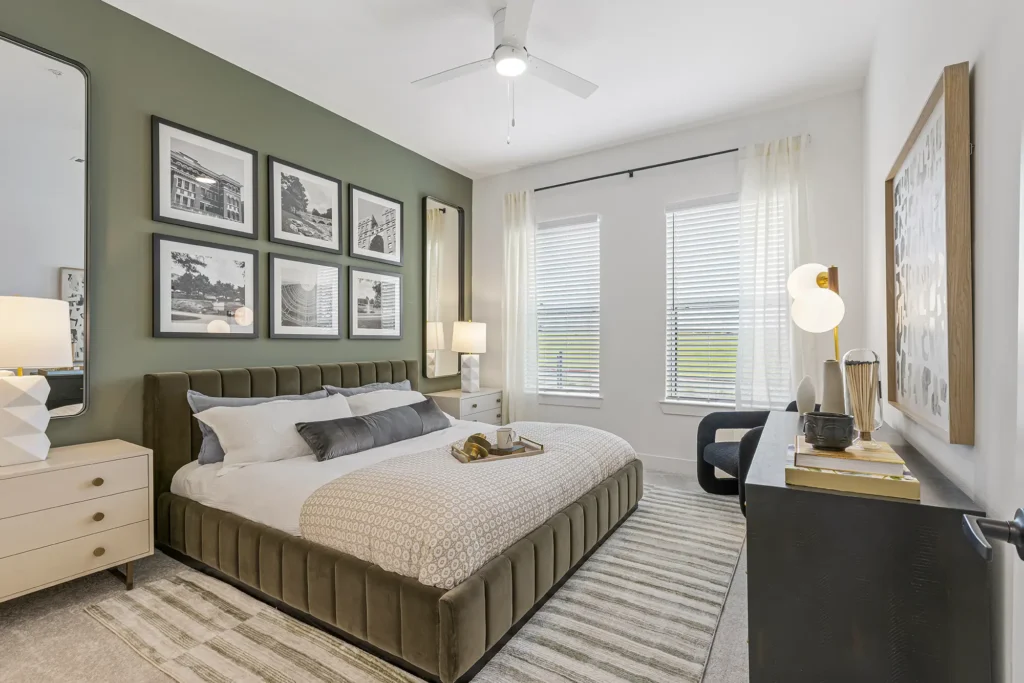 Modern bedroom with a green accent wall, large bed, neutral bedding, two nightstands with lamps, wall art above the bed, a rug, dresser, chair by the window, and natural light streaming through two large windows.