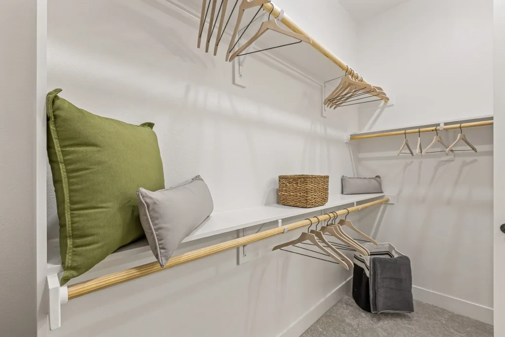 A walk-in closet with wooden hangers on two racks, a green pillow and a gray pillow on a white shelf, a wicker basket, and a gray fabric storage bin on the floor. The walls are white and the floor is carpeted.