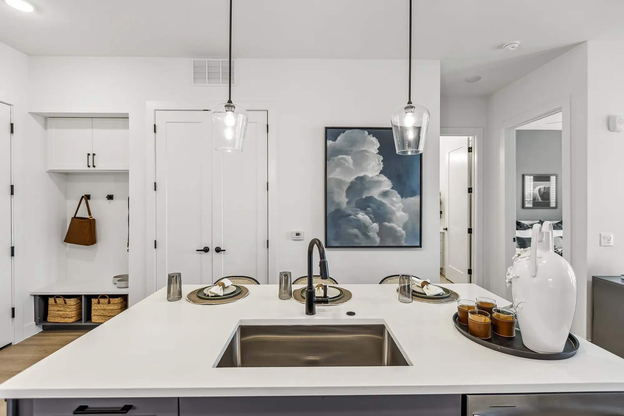 Modern kitchen with a large white island, built-in sink, and place settings for three. Pendant lights hang above, and a painting of clouds is on the back wall. Baskets and a coat are visible in a small nook to the left.