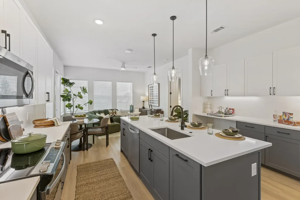 Modern kitchen with white and gray cabinets, a large island with sink, pendant lights, and place settings. Open layout connects to a living area with plants, green sofa, windows, and wooden floors.