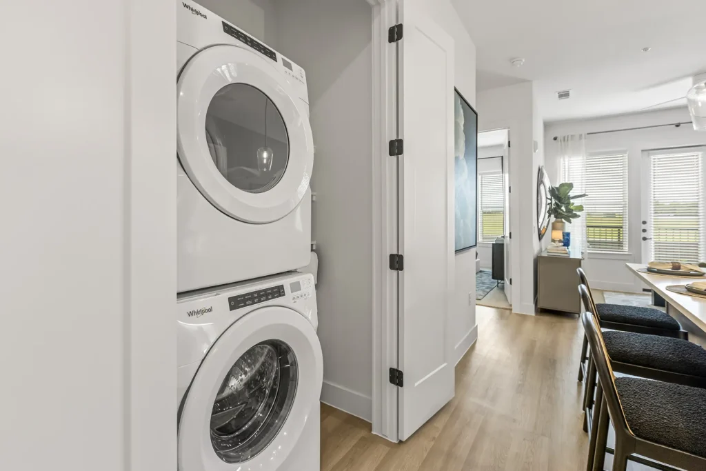 A modern laundry area with a stacked white washer and dryer unit is situated in a hallway next to an open door, leading to a bright dining and living space with large windows and wooden flooring.