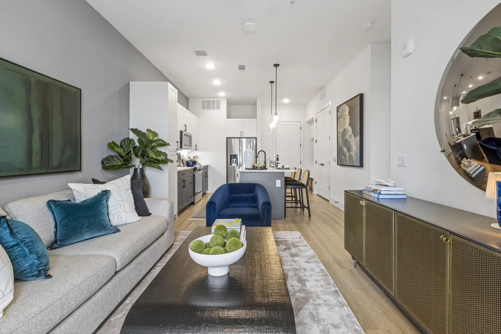 Modern open-concept living room and kitchen with neutral tones, a beige sofa with blue pillows, a blue armchair, a black coffee table, contemporary art, and sleek kitchen appliances.