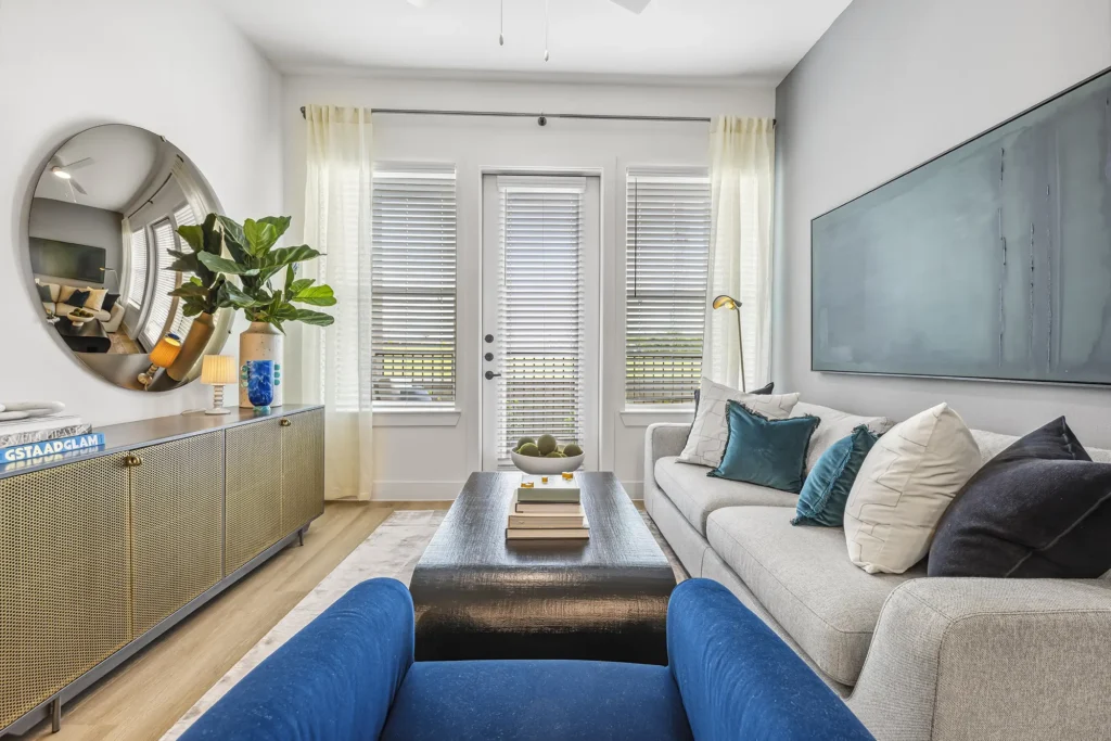 Modern living room with a gray sofa, blue armchair, coffee table, large abstract wall art, round mirror, sideboard with decor, potted plant, and French doors with white blinds and sheer curtains letting in natural light.
