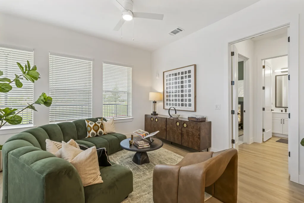 A modern living room with a green curved sofa, brown armchair, round coffee table, and mid-century cabinet. Large windows with blinds let in natural light. Decor includes throw pillows, a lamp, and wall art.