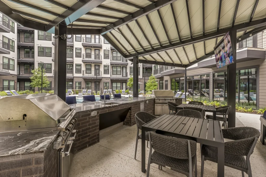 Outdoor covered patio area with a grill station, dining table, and chairs overlooking a modern apartment complex courtyard with pool and lounge chairs; TV mounted above the grilling area.