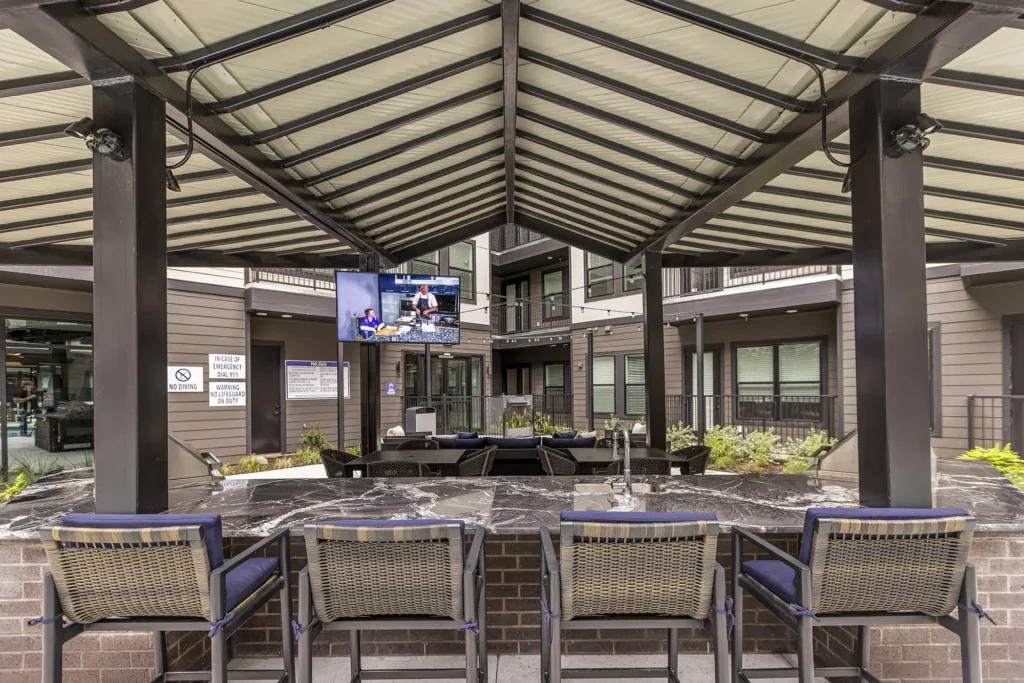 Outdoor covered patio area with a marble counter, bar stools, mounted TV screen, and lounge seating, surrounded by a modern apartment complex. The space has plants and a relaxed, social atmosphere.