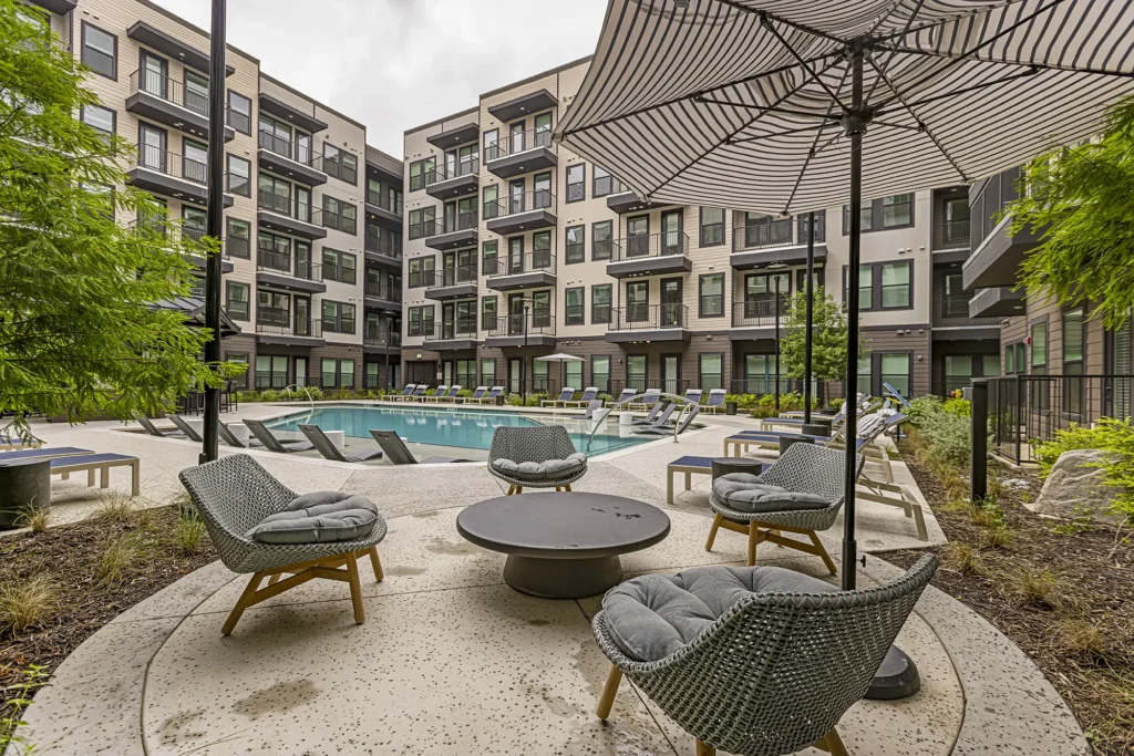 Modern apartment courtyard with a swimming pool, lounge chairs, a shaded seating area with cushioned chairs and a round table, surrounded by greenery and a four-story building in the background.