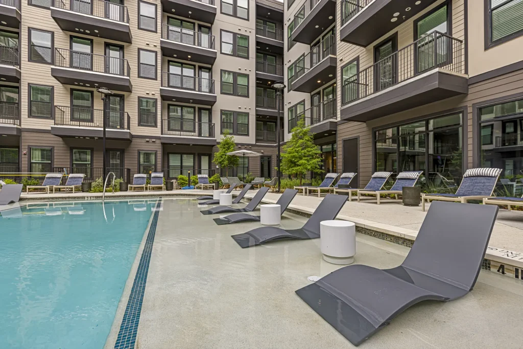 Modern apartment complex with a swimming pool, gray lounge chairs partially submerged in the water, small white tables, and additional loungers on the pool deck. Large glass windows and balconies overlook the pool area.
