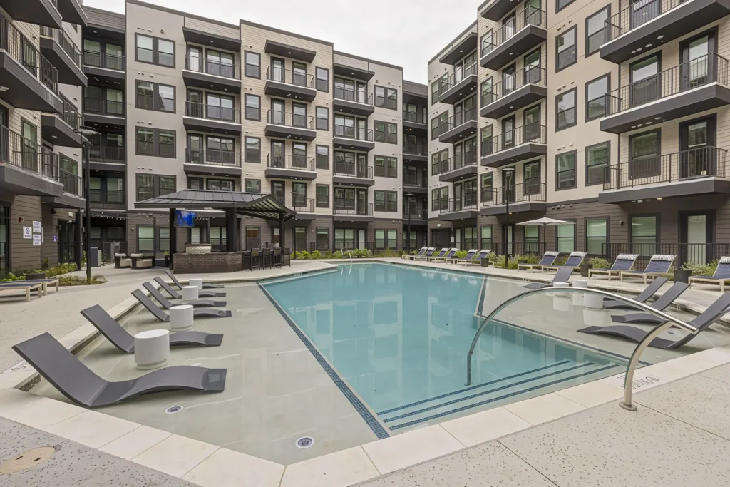 Modern apartment complex courtyard with a large outdoor swimming pool, lounge chairs partially submerged in the water, shaded seating area, and four-story buildings with balconies surrounding the pool.