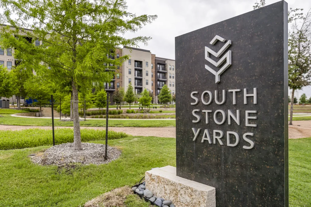 A large sign reading South Stone Yards stands in front of a landscaped area with trees and grass, with modern apartment buildings visible in the background under a cloudy sky.